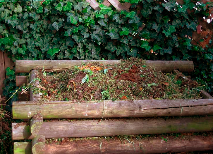 A timber compost bin with interlocking corners, stacked boards and garden material, against a leafy background.