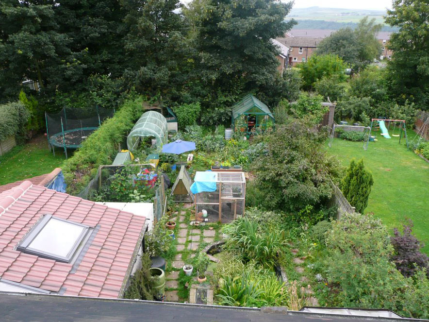 An elevated view of a suburban garden with paths, planted beds, small greenhouses and trees.